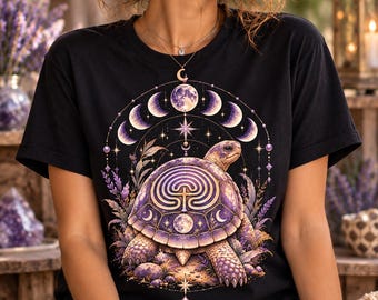 Può includere: Una t-shirt nera a maniche corte con una stampa grafica di una tartaruga con un motivo a labirinto sul guscio. Il design include elementi celesti viola e dorati, fasi lunari, stelle e rametti di lavanda che circondano la tartaruga.