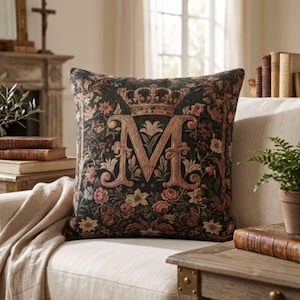 May include: A square throw cushion featuring a dark green tapestry fabric with a woven floral pattern in shades of pink, cream, and gold. The centre displays a large, ornate letter M topped with a detailed crown, all rendered in a golden thread.