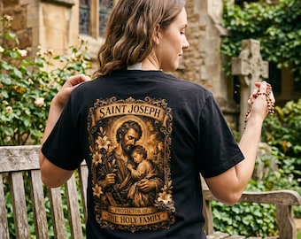 May include: A black t-shirt featuring a detailed graphic on the back. The print depicts Saint Joseph holding the child Jesus, surrounded by lilies, with the text "SAINT JOSEPH" above and "PROTECTOR OF THE HOLY FAMILY" below in a gold frame.