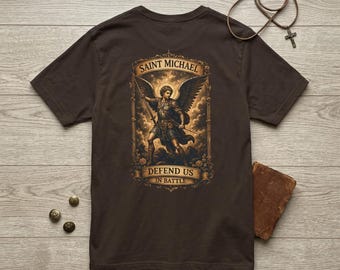 May include: A dark brown t-shirt featuring a graphic print of Saint Michael with wings holding a spear, framed by an ornate border. The text reads Saint Michael Defend Us In Battle. A brown leather book and a cross necklace are also present.