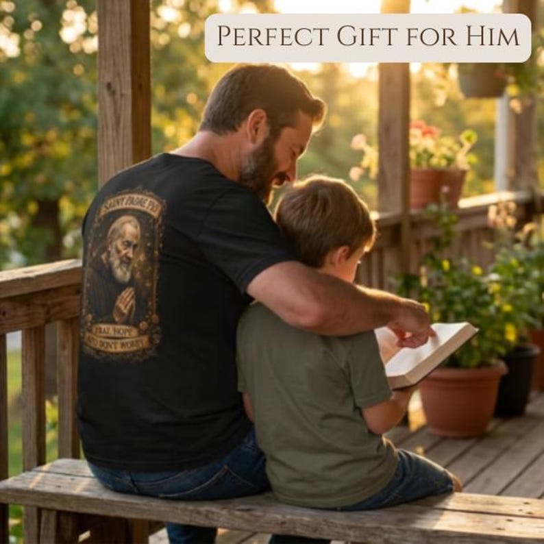 May include: A black t-shirt featuring a graphic print of Saint Padre Pio with the text "Pray, Hope and Don't Worry" enclosed in an ornate frame. The design is centered on the back of the shirt, which is worn by a person sitting on a wooden bench.