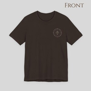 A dark brown short-sleeve t-shirt featuring a circular graphic on the left chest. The graphic contains a cross symbol surrounded by text and a dotted border.