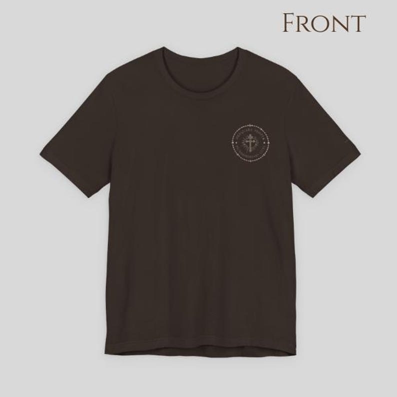 A dark brown short-sleeve t-shirt featuring a circular graphic on the left chest. The graphic contains a cross symbol surrounded by text and a dotted border.