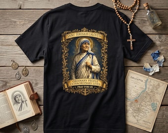 May include: A black t-shirt featuring a central graphic of Saint Teresa of Calcutta within an ornate gold frame. The text reads: SAINT TERESA OF CALCUTA, ORDINARY PEOPLE EXTRAORDINARY LIVES, PRAY FOR US. The design includes a halo and rosary beads.
