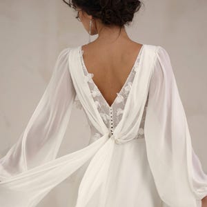 Puede incluir: Vestido de novia blanco con espalda en V baja y mangas largas y fluidas. La espalda presenta delicado encaje floral y detalles de botones. El vestido tiene una falda fluida y la modelo sostiene la tela.