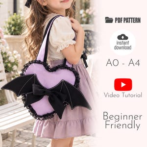 Puede incluir: Un bolso en forma de corazón de color lavanda con alas de murciélago negras, un lazo negro y ribete de encaje negro. La imagen incluye el texto: PDF PATTERN, instant download, A0 - A4, Video Tutorial y Beginner Friendly. Bolso con dos asas.