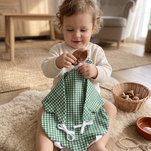 May include: A green and white gingham patterned baby romper featuring a white collar, short sleeves, and two white fabric ties on the front. The garment is made of a lightweight textile and includes a small brown fabric detail near the neckline.