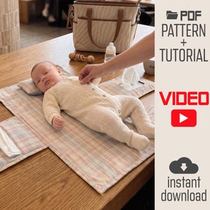 May include: A baby lies on a plaid fabric changing mat with a small pillow. The text reads PDF PATTERN + TUTORIAL, VIDEO, and instant download. A beige tote bag, a baby bottle, and a tissue dispenser are visible in the background.
