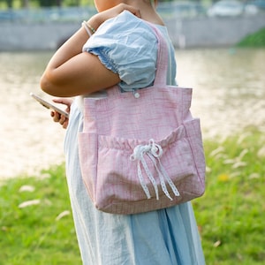 May include: A light pink tote bag with a subtle textured pattern, featuring two top handles, a snap button closure, and a front exterior pocket with an elasticized opening decorated with a white lace drawstring bow detail.