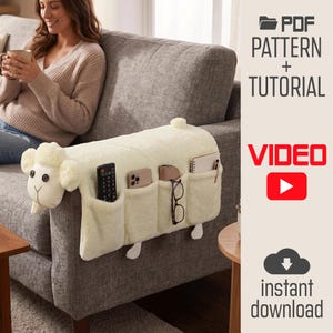 Puede incluir: Un organizador de reposabrazos de sofá de felpa color crema con forma de oveja, con cabeza de ojos de botón negros, cuatro bolsillos con mando, smartphone, gafas y cuaderno, y el texto PDF PATTERN + TUTORIAL VIDEO.