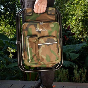 May include: A camouflage print folding stool with an integrated backpack featuring the name Martin embroidered in white script. The stool has a black metal frame, a main zippered compartment, a side pocket, and a front pocket with a silver trim.
