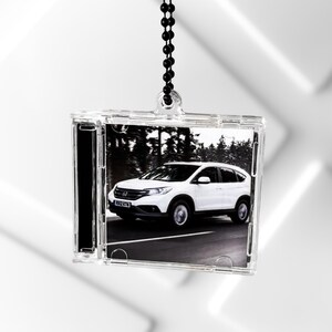 Könnte beinhalten: Ein quadratisches Ornament aus klarem Kunststoff mit einem Schwarz-Weiß-Foto eines weißen Honda CR-V, der auf einer Straße fährt. Das Ornament ist an einer schwarzen Kugelkette aufgehängt, die an einer kleinen Öse oben am Gehäuse befestigt ist.