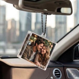 Könnte beinhalten: Ein quadratischer, durchsichtiger Auto-Hängeanhänger aus Acryl, der an einer schwarzen Kugelkette befestigt ist. Der Anhänger zeigt ein aufgedrucktes Foto eines Paares, das sich anlächelt, in einem transparenten Rahmen am Rückspiegel.