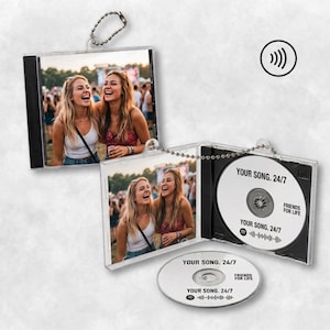 Könnte beinhalten: Ein personalisierter CD-Schlüsselanhänger mit einer transparenten Kunststoffhülle, die ein Foto von zwei lachenden Personen bei einer Outdoor-Veranstaltung zeigt. Die Disc im Inneren und die lose Disc tragen den Text "YOUR SONG. 24/7" und "FRIENDS FOR LIFE".