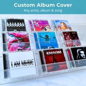 Könnte beinhalten: Ein mehrstufiger Präsentationsständer aus klarem Acryl mit mehreren quadratischen CD-Hüllen und verschiedenen Albumcovern, darunter "I AM MUSIC" und "The Eminem Show". Das Regal hat drei horizontale Ebenen mit verschiedenen Musik-CDs.
