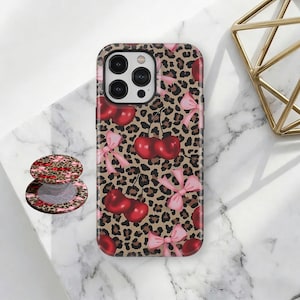 Op de afbeelding: Een telefoonhoesje met een bruine luipaardprint, versierd met rode kersen en roze strikken. Een bijpassende telefoonhouder is ook zichtbaar. De hoes heeft een glanzende afwerking en een beschermend ontwerp.