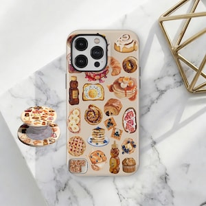 Może przedstawiać: Beżowe etui na telefon i pasujący popsocket z akwarelową ilustracją różnych wypieków śniadaniowych. Projekt obejmuje naleśniki, rogaliki, pączki i inne wypieki. Etui na telefon ma wycięcie na aparat.