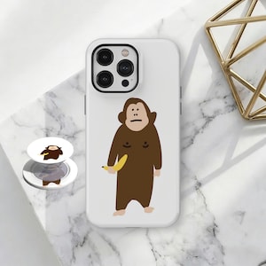 May include: White phone case with a cartoon monkey holding a banana. A matching phone grip is also visible. The monkey is brown with a simple facial expression. The phone case has a black camera lens surround.