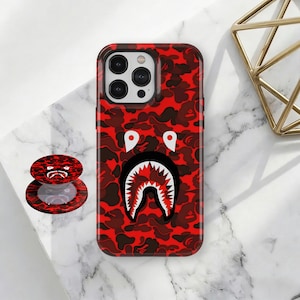 May include: Red camouflage phone case with a shark face design. A matching pop-up phone grip is also visible. The case and grip feature a red and brown camouflage pattern with a white and black shark face.