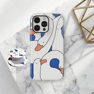 Könnte beinhalten: Weißes Handycase mit einem Muster aus Cartoon-Gänsen mit orangefarbenen Schnäbeln und blauen Akzenten. Ein passender Handyhalter ist ebenfalls sichtbar. Das Handycase liegt auf einer Marmoroberfläche.