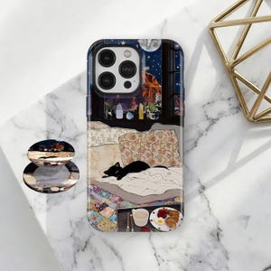 May include: A phone case depicting a bedroom scene with a black cat on a bed. The case has a dark blue background with a window view of a moonlit night and a fireplace. A matching pop socket is also visible.