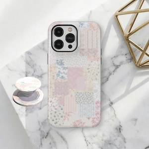 Pode incluir: Uma capa de telefone e um PopSocket a condizer com um design de patchwork em cores pastel. A capa apresenta uma mistura de tecidos florais e estampados em rosa, azul e branco. O PopSocket tem um design semelhante.