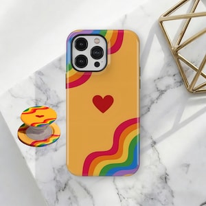 May include: A yellow phone case featuring a rainbow design and a red heart. A matching rainbow PopSocket is next to the phone. The case and PopSocket have a colourful, cheerful design.