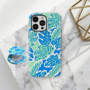 May include: A smartphone case and a matching circular collapsible phone grip featuring a vibrant botanical pattern of overlapping monstera and fern leaves in shades of bright blue, light blue, and pale mint green against a white background.