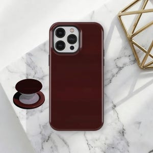 May include: A burgundy phone case with a matching pop-up grip. The phone case has a glossy finish and a camera cutout. The pop-up grip is round and has a dark burgundy top with a white base. A gold geometric decoration is in the background.