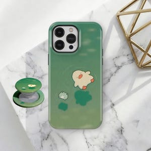 May include: Green phone case with a cartoon duck and frog design. A matching green pop socket with a lily pad design is next to the phone case. The phone case has a gradient green color.