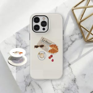Może przedstawiać: Białe etui na iPhone'a z nadrukowaną ilustracją gazety, rogalików, kawy, wiśni i okularów przeciwsłonecznych. Biały uchwyt pop socket z małą ilustracją znajduje się obok telefonu. Złota geometryczna dekoracja znajduje się w tle.