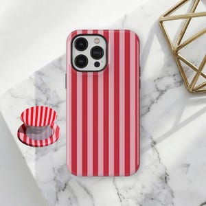 Puede incluir: Una funda de teléfono y un agarre a juego con un patrón de rayas rojas y rosas. La funda del teléfono está sobre una superficie de mármol blanco, con una decoración geométrica dorada en el fondo.