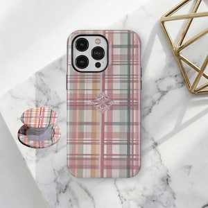 May include: A rectangular phone case featuring a plaid pattern in shades of pink, sage green, and tan, with a central decorative filigree emblem. Beside the case is a matching circular collapsible phone grip with the same plaid design.