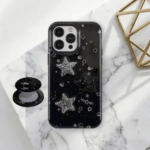 Puede incluir: Funda de teléfono negra con un diseño de estrellas brillantes. La funda presenta tres grandes estrellas plateadas con purpurina y pequeños detalles de estrellas. También se ve un pop socket negro y plateado a juego.