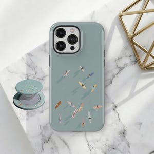 Puede incluir: Una funda de teléfono verde azulado con un diseño de surfistas en tablas de surf. La funda se combina con un pop socket verde azulado a juego con un diseño de escena de playa. Una decoración geométrica dorada está en el fondo.