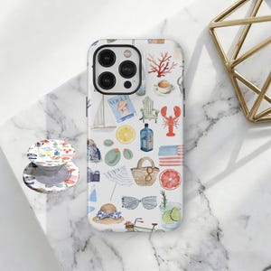 May include: White iPhone case with a nautical-themed pattern featuring a sailboat, lobster, sunglasses, and a miniature American flag. A matching PopSocket is visible. The case has a black camera lens border.