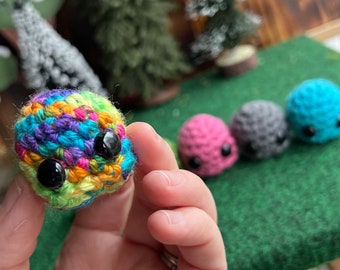 Könnte beinhalten: Nahaufnahme eines bunten, gehäkelten Balls mit großen schwarzen Augen. Der Ball wird in einer Hand gehalten, mit drei weiteren gehäkelten Bällen in Rosa, Grau und Blau im Hintergrund. Der bunte Ball hat eine Mischung aus Regenbogenfarben.
