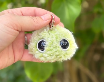 Könnte beinhalten: Ein flauschiger, hellgrüner Schlüsselanhänger mit großen, schwarzen, reflektierenden Augen. Er hat einen silberfarbenen Verschluss zum einfachen Anbringen an Schlüsseln oder Taschen. Das Plüschmaterial verleiht ihm ein weiches, taktiles Aussehen.