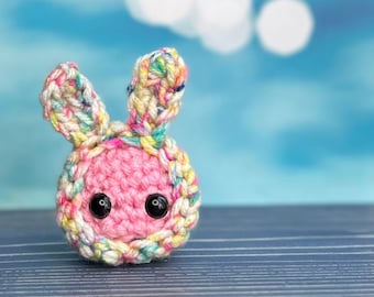 Könnte beinhalten: Ein kleiner rosa gehäkelter Hase mit bunten Garn-Akzenten und schwarzen Knopfaugen.