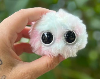 Könnte beinhalten: Ein kleines, flauschiges Plüschtier mit großen, schwarzen, glänzenden Augen. Das Spielzeug ist eine Mischung aus rosa, weiß und hellblauen Farben. Das Plüschtier wird in einer Hand vor einem verschwommenen grünen Hintergrund gehalten.