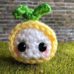 May include: A crocheted, round, yellow and white plush toy with black bead eyes and pink cheeks. The toy has a green crocheted leaf on top. The background is a brick wall and green surface.
