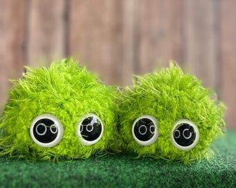 Könnte beinhalten: Zwei flauschige, lindgrüne Kreaturen mit großen, runden Augen mit weißem Rand und schwarzen Pupillen. Die Kreaturen bestehen aus einem weichen, strukturierten Material und sitzen auf einer grünen Oberfläche mit verschwommenem Holzgrund.