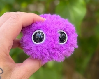 Könnte beinhalten: Ein flauschiges, lila Wesen mit großen, schwarzen Wackelaugen. Das Plüschtier hat eine runde Form und wird in einer Hand vor einem verschwommenen grünen Hintergrund gehalten. Ein skurriles, handgefertigtes Objekt.
