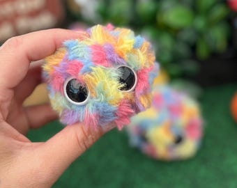 Könnte beinhalten: Ein flauschiges, rundes Plüschtier mit einem bunten Mix aus rosa, gelbem, blauem und lila Fell. Das Spielzeug hat große, schwarze Wackelaugen. Ein zweites, ähnliches Spielzeug ist im Hintergrund leicht unscharf.
