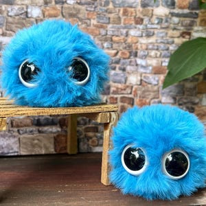 May include: Two fluffy, blue, round plush toys with large, black and white eyes. One toy sits on a small wooden table, while the other rests on a wooden surface. The toys have a shaggy texture.