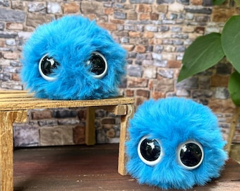 Könnte beinhalten: Zwei flauschige, blaue, runde Plüschtiere mit großen, schwarz-weißen Augen. Ein Spielzeug sitzt auf einem kleinen Holztisch, während das andere auf einer Holzoberfläche liegt. Die Spielzeuge haben eine zottelige Textur.