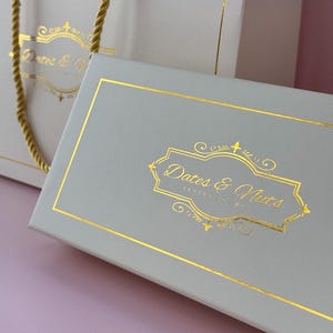 May include: A light grey gift box and matching shopping bag with gold trim and handles. The box and bag feature the text "Dates & Nuts" in gold script within an ornate gold frame.