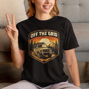 May include: Black t-shirt with a graphic featuring an off-road vehicle, mountains, and the text "OFF THE GRID." The design is in a vintage style with a sunset background. The shirt is made of a soft material.