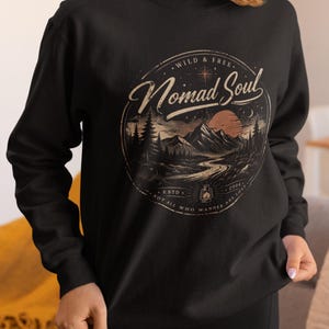 Könnte beinhalten: Schwarzes Sweatshirt mit einem kreisförmigen Grafikdesign, das eine Berglandschaft, einen Fluss und einen Sonnenuntergang zeigt. Das Design enthält den Text "Nomad Soul", "Wild & Free" und "Not All Who Wander Are Lost".