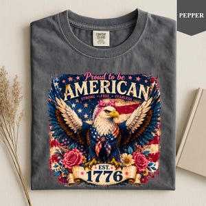 May include: Dark grey t-shirt featuring a patriotic design. The graphic includes an American flag, an eagle, and the text "Proud to be AMERICAN" with "Strong, Free, Fearless" below. The year "1776" is also displayed.